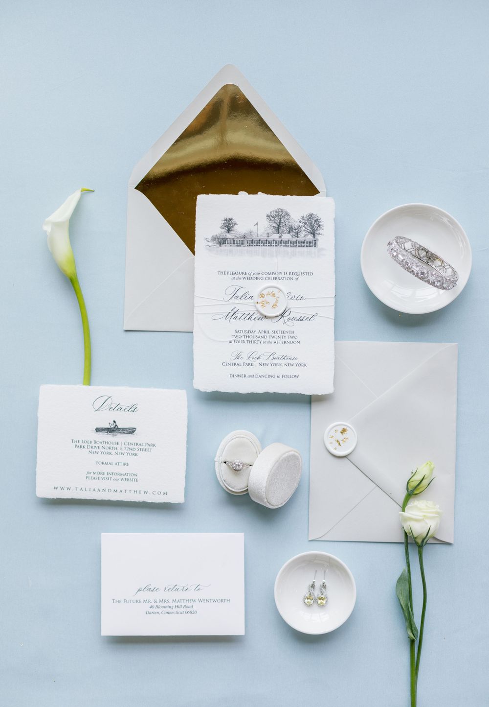 Wedding invitation suite with white flowers, gold envelope, and delicate styling details on light blue background.