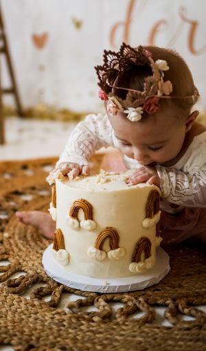 Small child in white dress leaning over white cake decorated with gold rainbows.