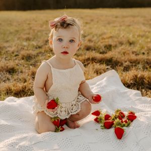 Toddler in lace dress sitting on white blanket with fresh strawberries scattered around.