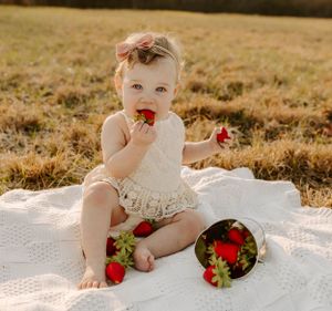 Sweet baby in white dress eating strawberries during outdoor photoshoot on white blanket.