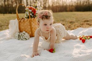Baby in white dress crawling on blanket with picnic basket and flowers in golden field.
