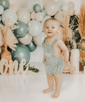 A one year old in a gray romper smiles while standing against a balloon backdrop with sage and white balloons.