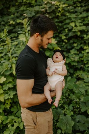 Father holds smiling baby in white onesie against green leafy background.