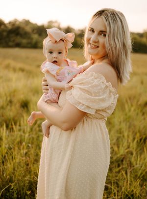 Mother holds baby in matching cream dresses standing in sunlit meadow.