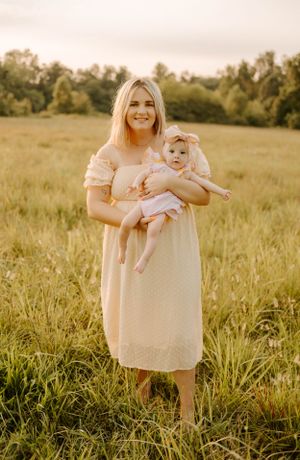 Mother and baby in matching cream dresses pose together in golden field at sunset.