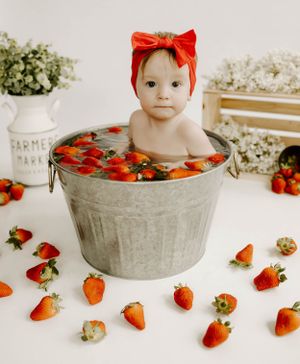 A creative baby photoshoot with fresh strawberries scattered around a galvanized metal tub.