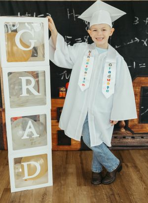 A graduate in white cap and gown poses next to alphabet blocks spelling GRAD against a chalkboard.