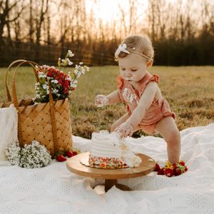Baby in pink romper reaching for birthday cake during sunset photoshoot outdoors.