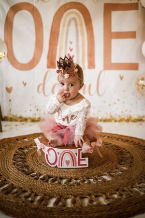 First birthday photoshoot with small crown, lace dress and tutu on round woven rug with gold lettering backdrop.