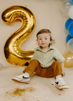 Toddler in casual outfit sitting by gold number 2 balloon with blue and white decorations.