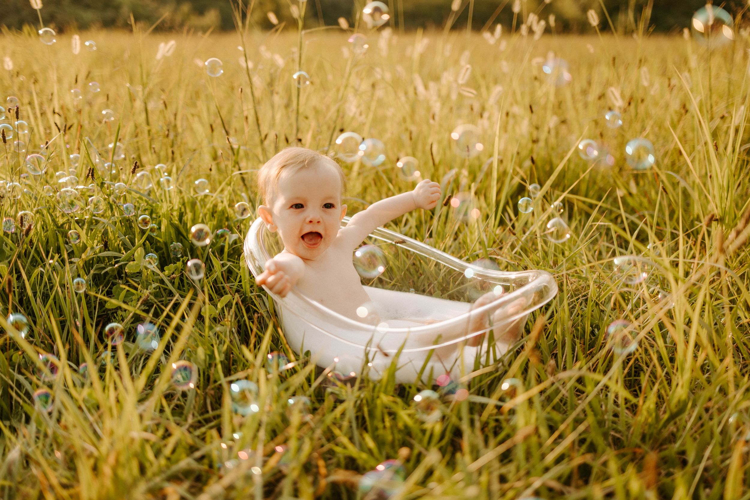Baby in white dress sits in small bathtub surrounded by tall grass and dandelions at sunset.