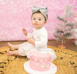 Baby in white outfit with bow having cake smash photo session against pink sparkly background.