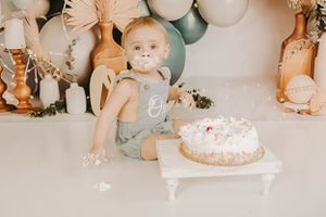 A first birthday celebration setup with white and sage balloons, pampas grass, and a frosted cake.