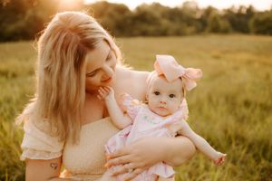 Mother cradles baby in cream dresses against golden sunset in field.