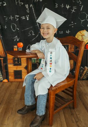 A graduate sits in wooden chair wearing decorated cap and gown against chalkboard with math equations.