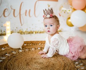 Baby in pink tutu and crown sitting on woven rug with balloon decorations and gold lettering backdrop.