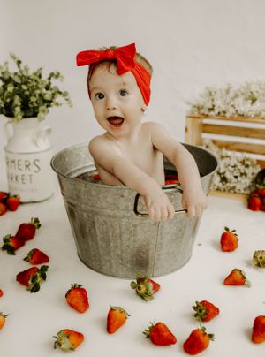 An excited baby in a red headband poses inside a vintage metal bucket with scattered strawberries.