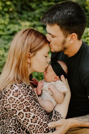 Parents share tender moment with newborn baby against leafy background.