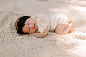 Sleeping baby in white lace outfit rests on textured cream blanket.