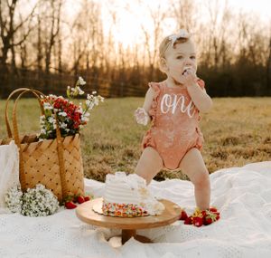 Baby wearing pink one outfit celebrating first birthday with cake outdoors.