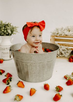 A joyful baby wearing a red bow sits in a metal bucket surrounded by fresh strawberries.