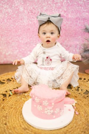 Baby in white lace dress and bow sitting with pink birthday cake against sparkly pink backdrop.