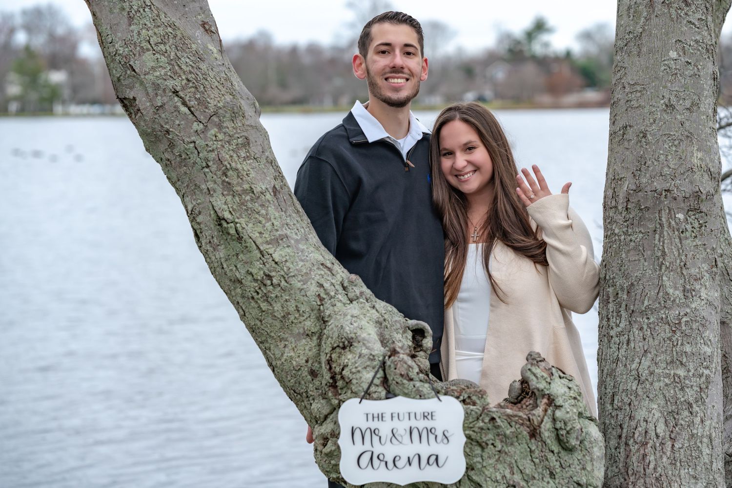 A newly engaged couple poses by a tree with a decorative sign.