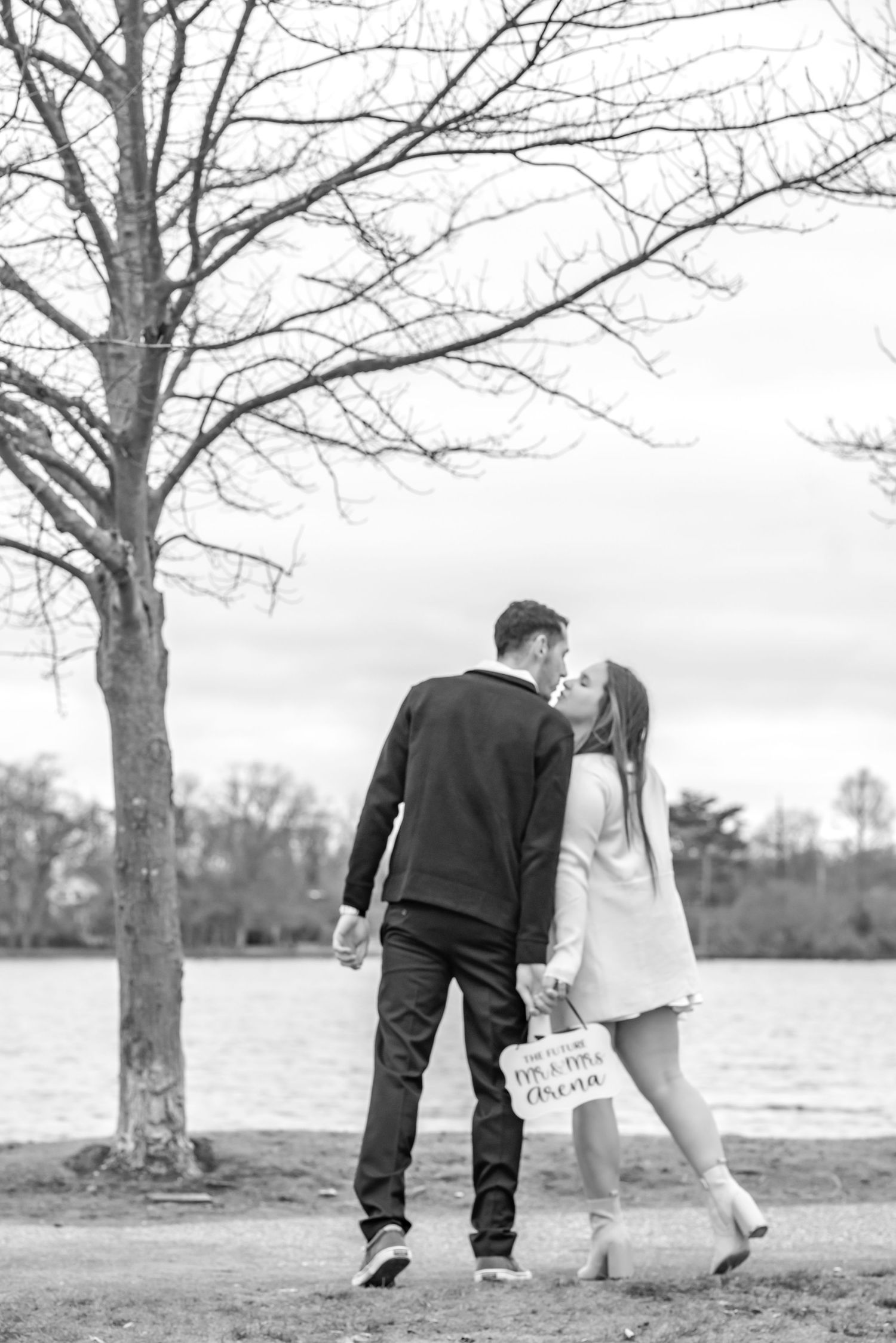A romantic black and white photo of a couple holding a sign by a lake.