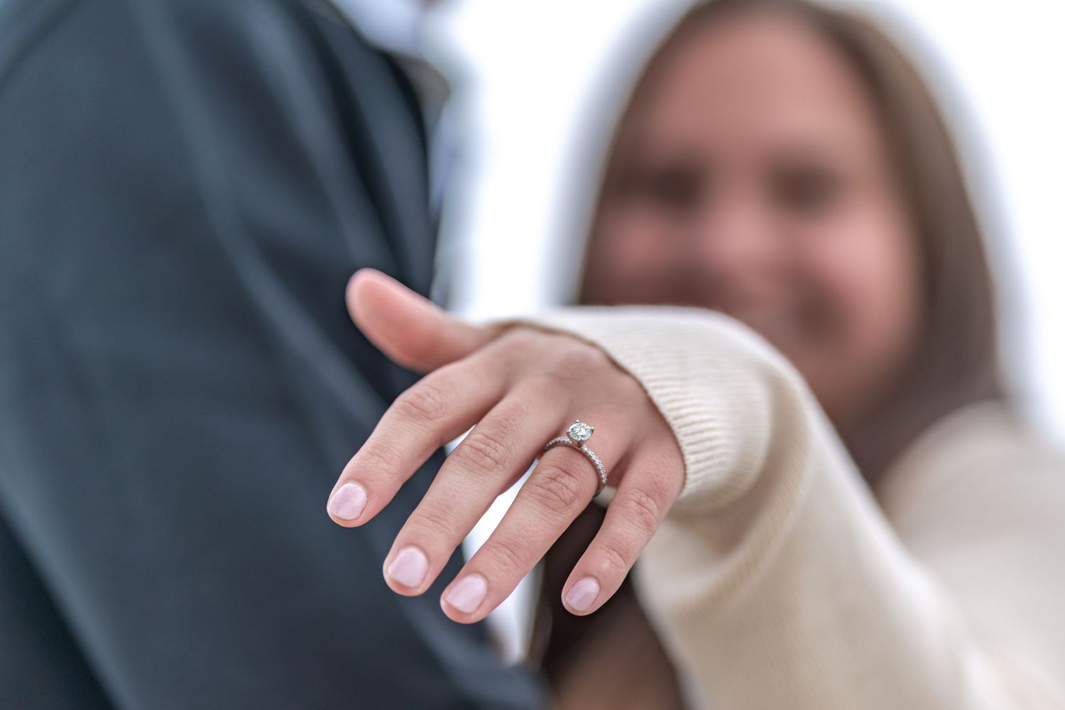 Close-up shot of a hand displaying an engagement ring against a dark jacket.