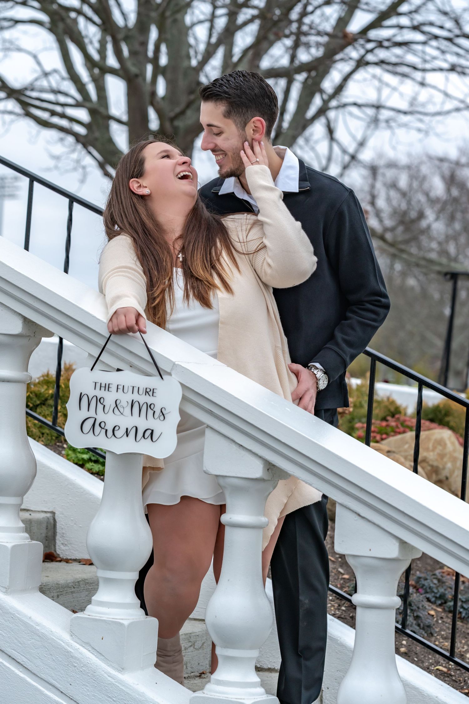 A couple shares a candid laughing moment on white stairs with a decorative sign.