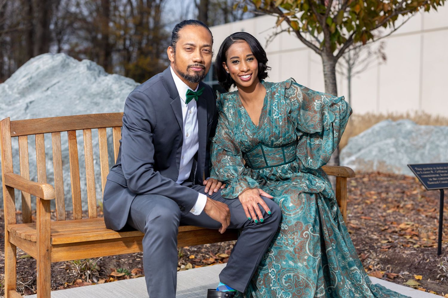 A couple sits together on a wooden bench outdoors, dressed in formal attire with a teal dress and grey suit.