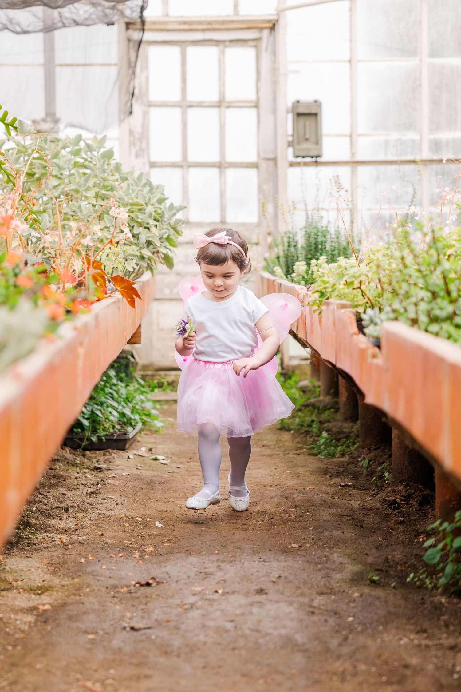 A small figure in a pink tutu and white dress walks between raised garden beds in a greenhouse setting.