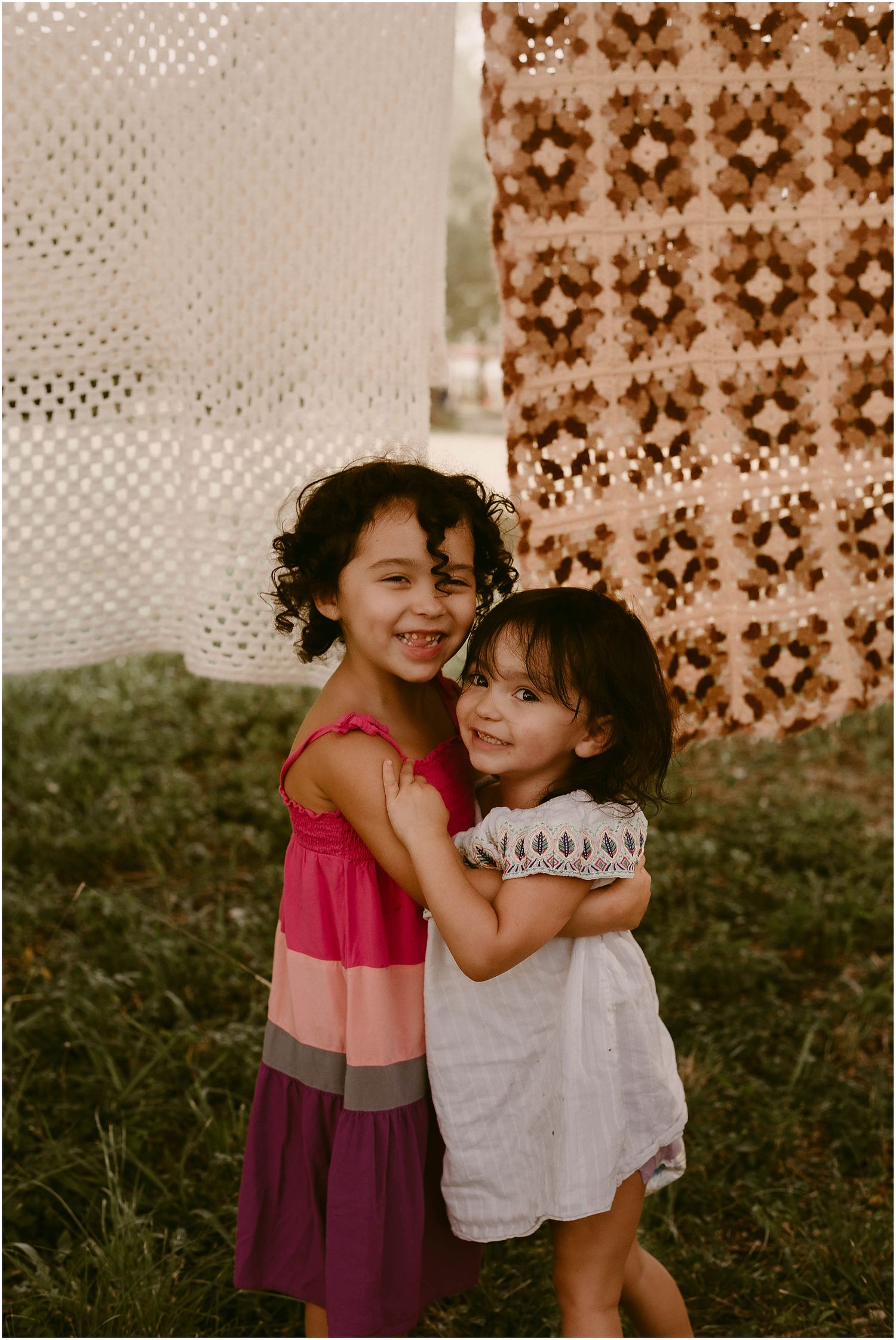 Two children embracing in front of patterned tapestries hanging outdoors.