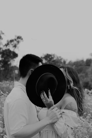 A black and white portrait of a couple sharing a hidden kiss behind a wide-brim hat.
