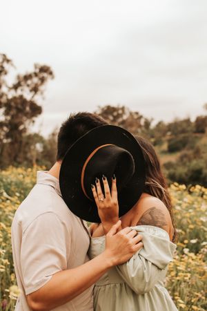 A romantic couple shares an intimate moment behind a black hat in a wildflower field.