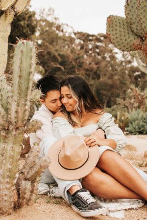 Romantic couple embracing near desert cactus while sitting with a tan hat.