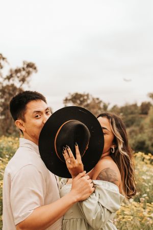 Playful couple hiding behind a black hat in a field of yellow flowers.
