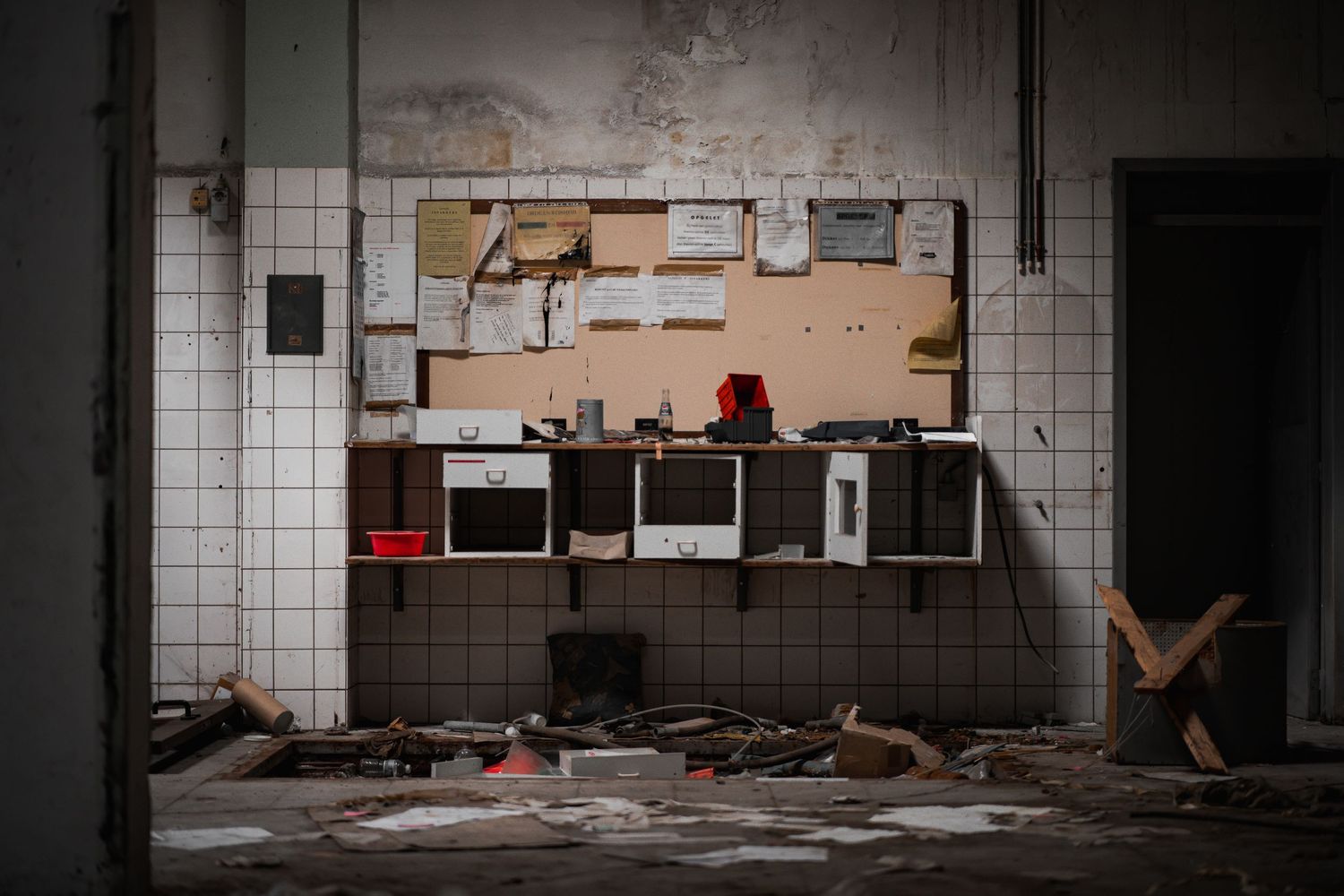 An abandoned industrial kitchen with damaged white tile walls and decaying appliances in a dark, deteriorating space.