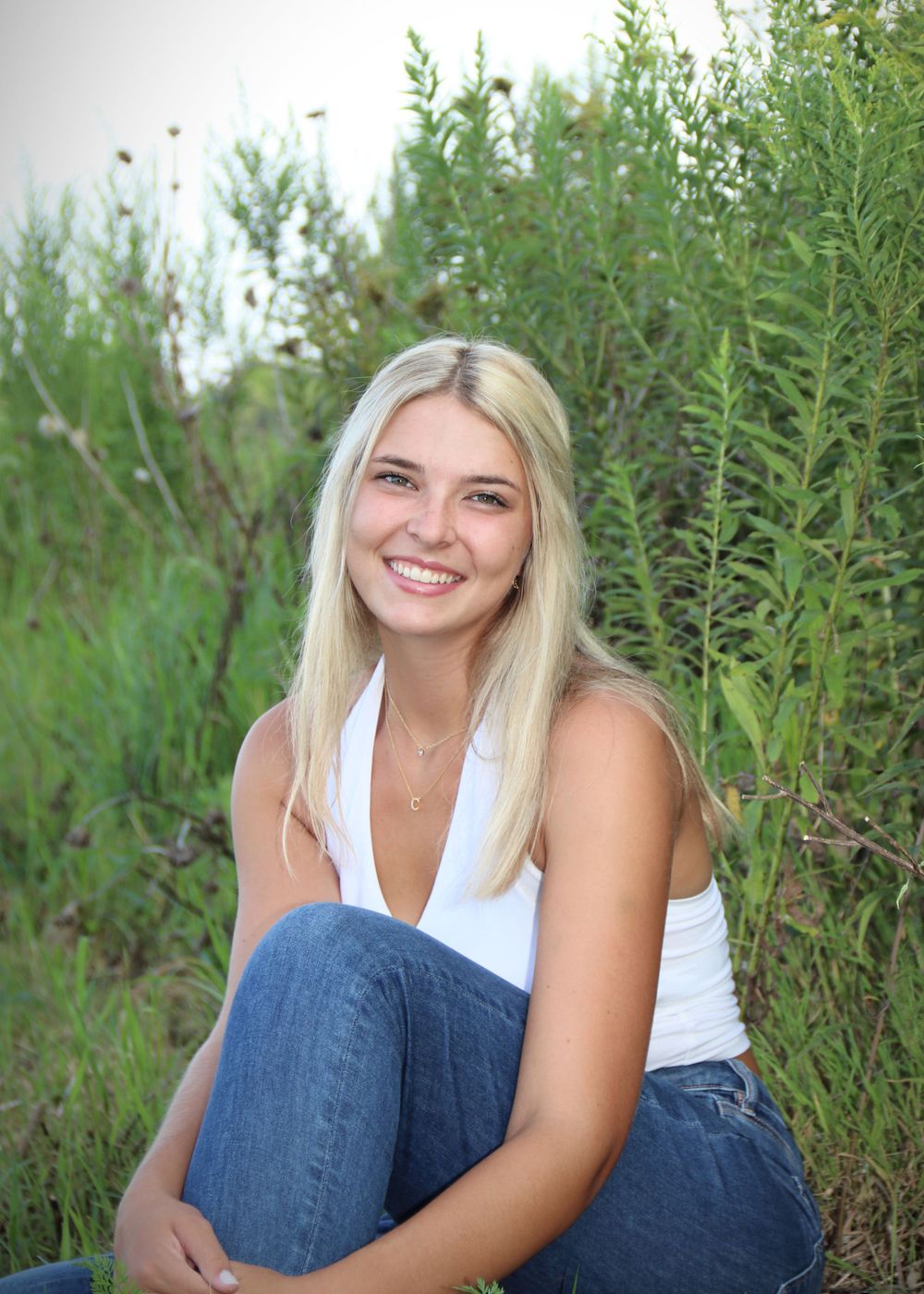 Outdoor portrait in white tank top and jeans surrounded by tall green plants.
