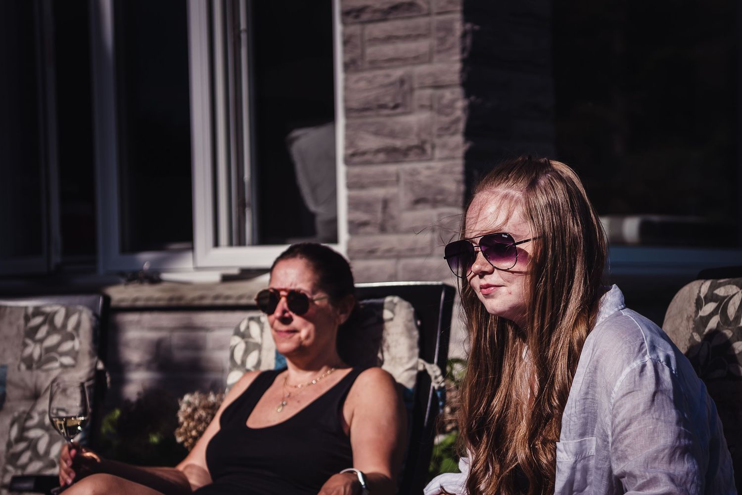 Colour image of a woman and her teenage daughter sitting outside on deck chairs and wearing sun glasses.