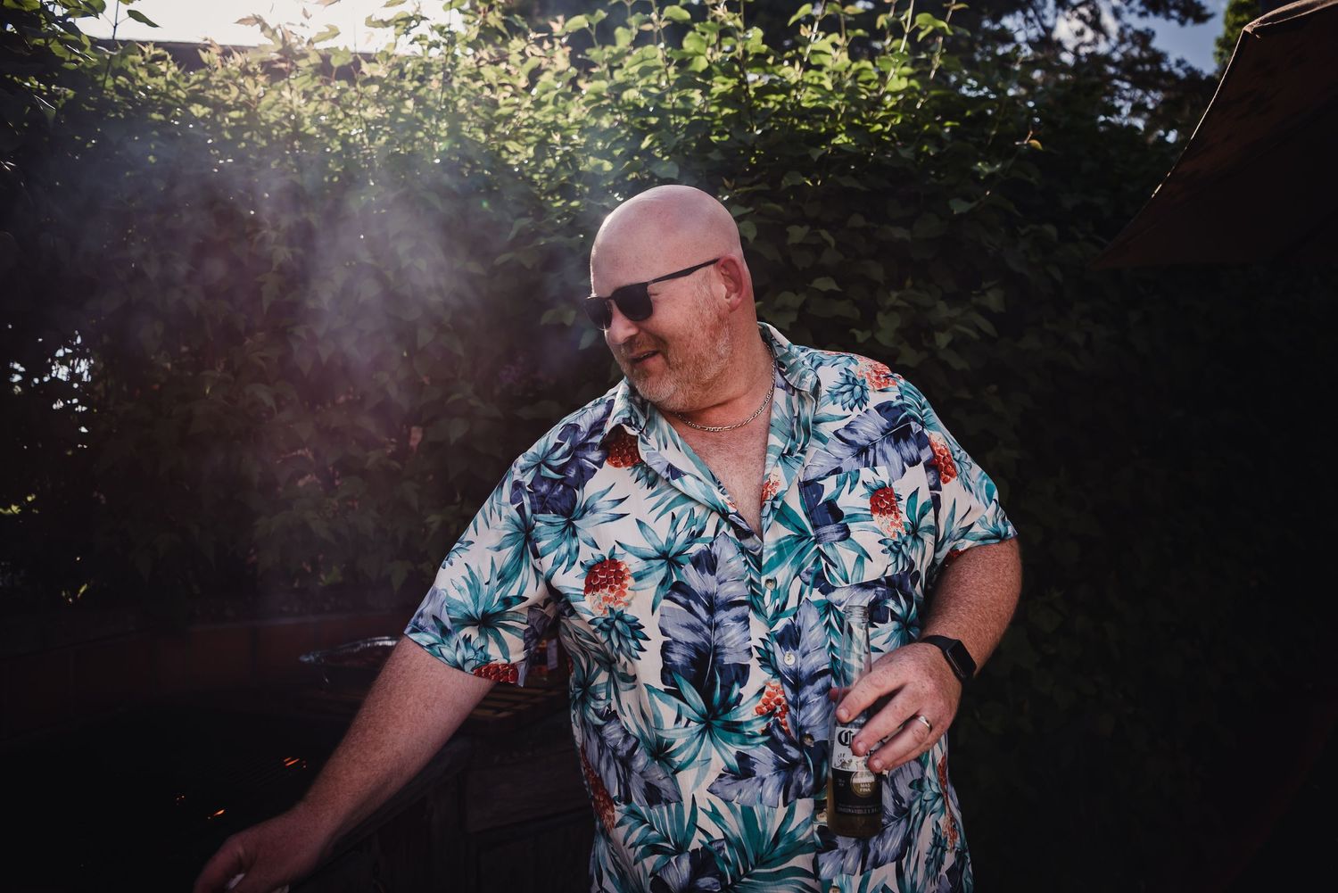 Colour image of a caucasian man bbqing as the sun shines over the bushes behind him.