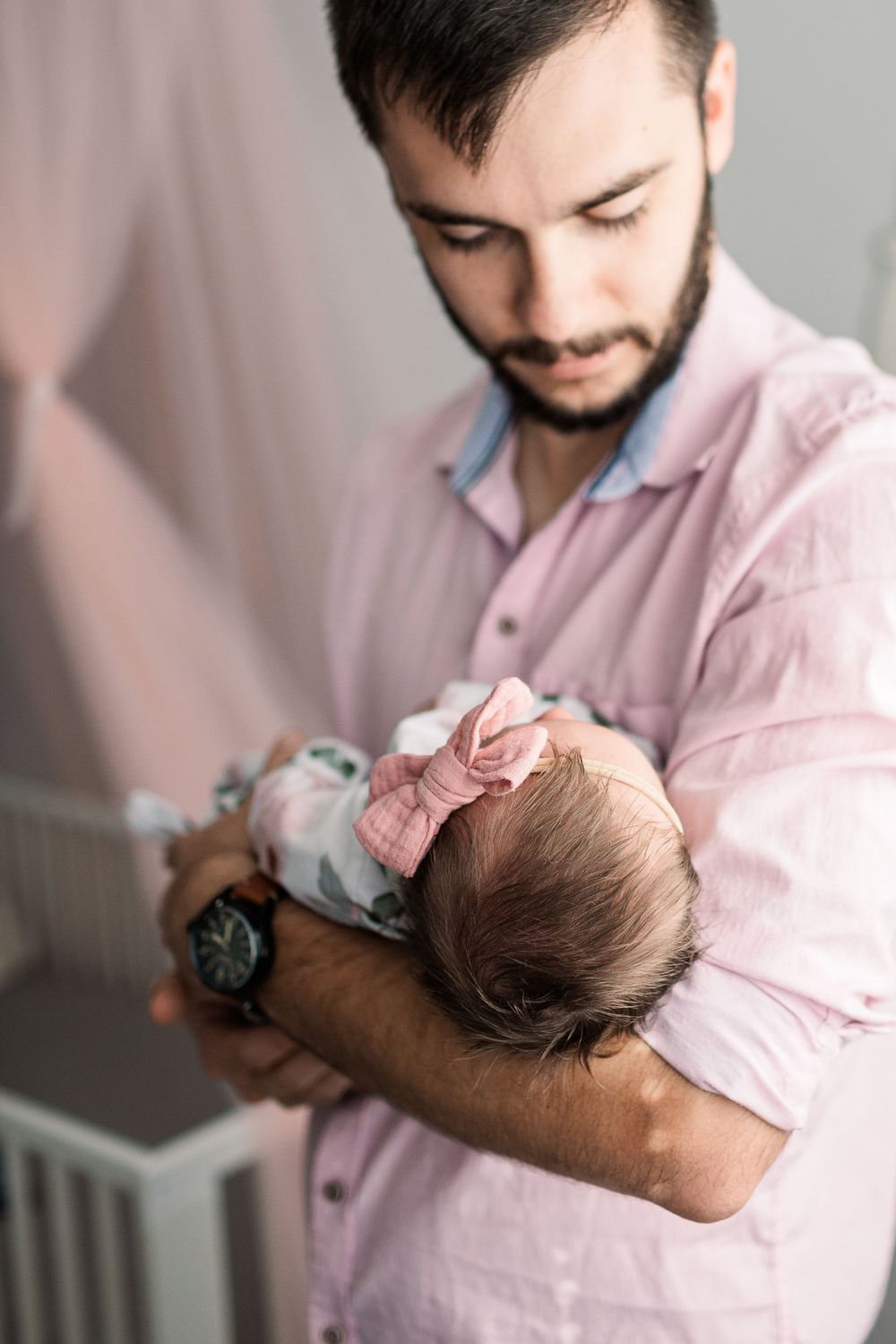 Father in pink button-down shirt gently holding newborn baby wearing floral outfit.