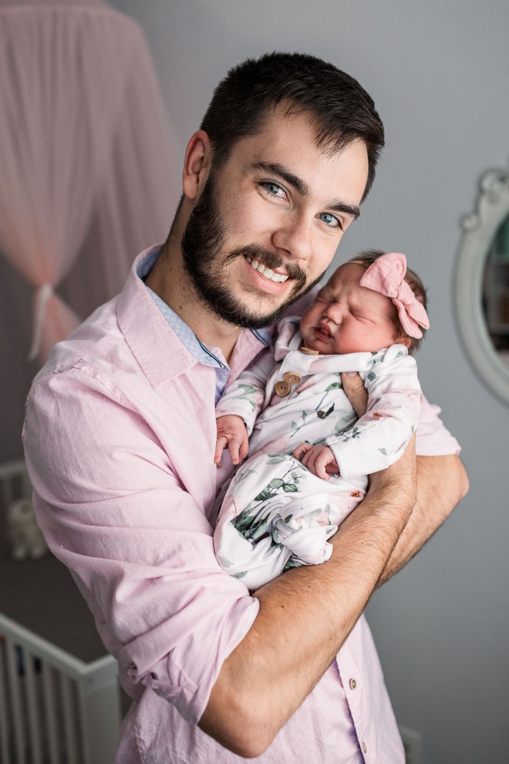 Father in pink shirt holding sleeping newborn wrapped in floral swaddle blanket.