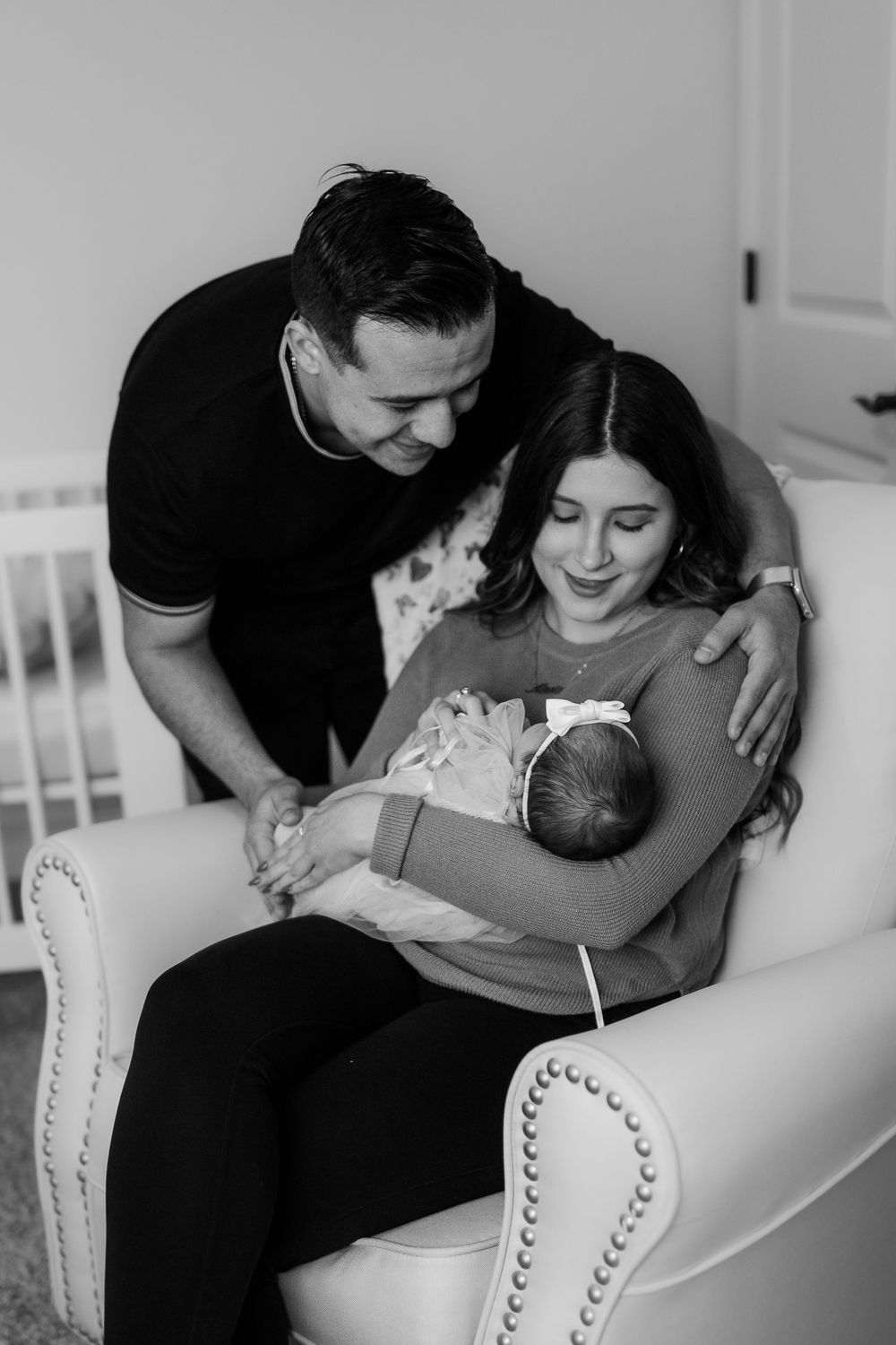 Black and white photo of parents sharing tender moment with newborn in nursery chair.