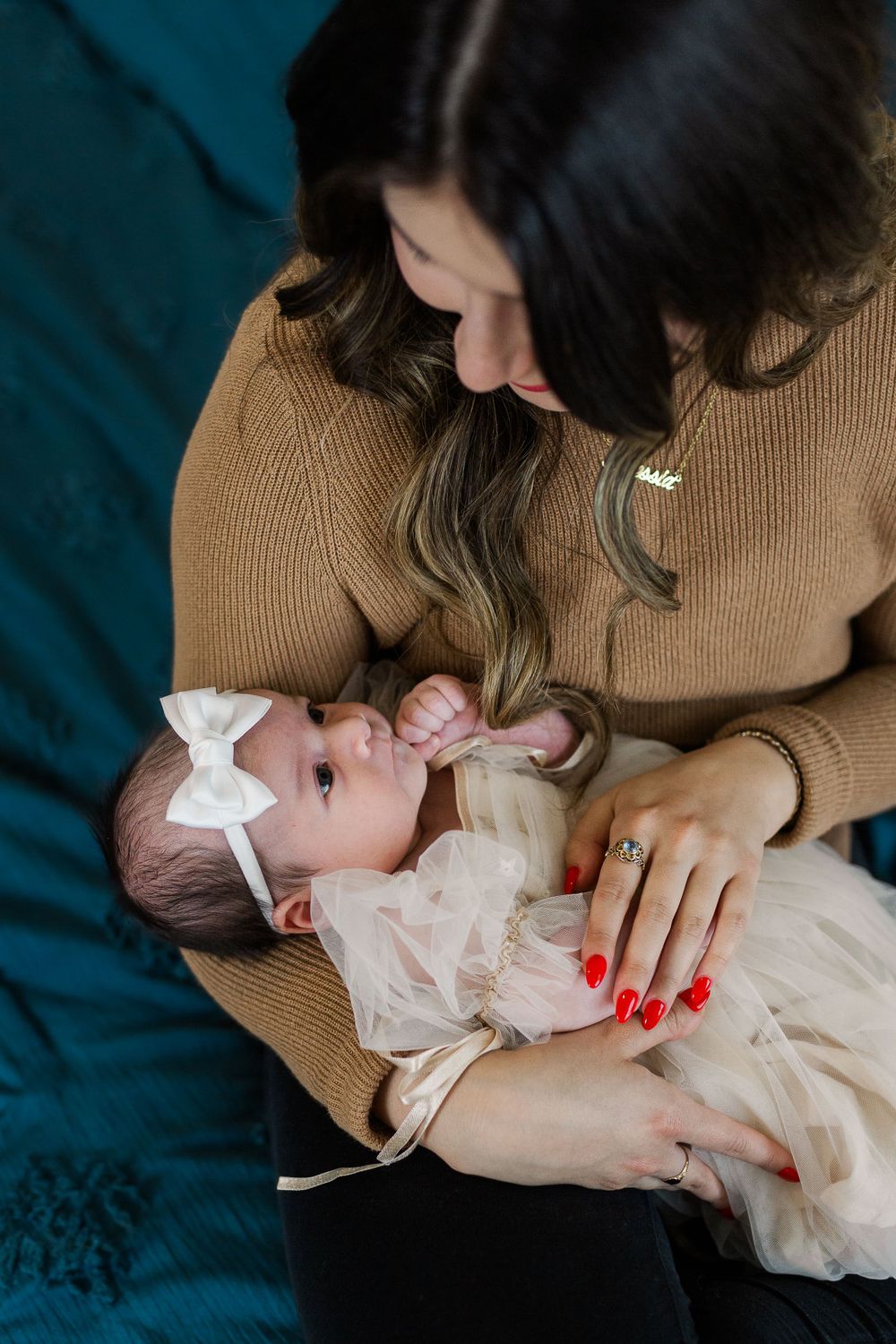 Mother in brown sweater gazing lovingly at baby in white dress with bow.