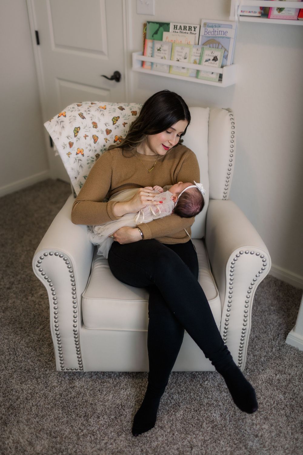 Mother in brown sweater holding sleeping newborn in white nursery chair.