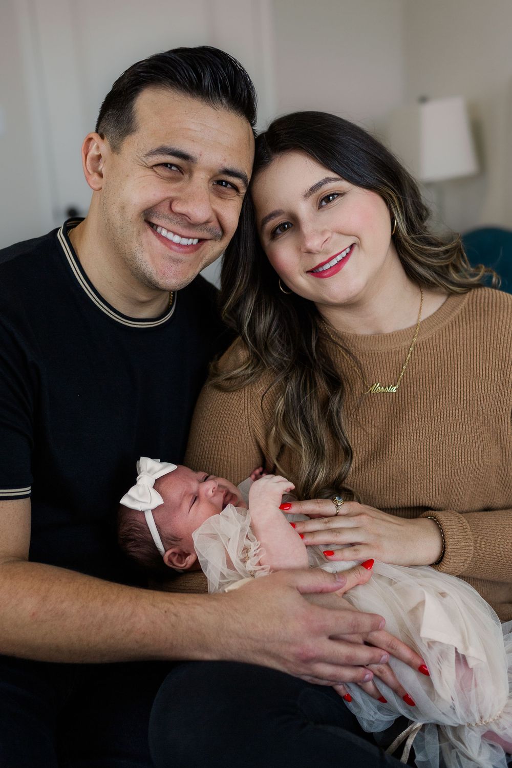 Parents in casual attire sharing happy moment with newborn in white dress.