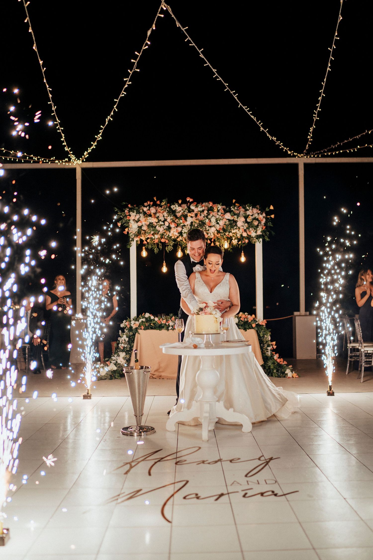 Wedding cake cutting scene with sparklers and floral chandelier creating magical atmosphere.