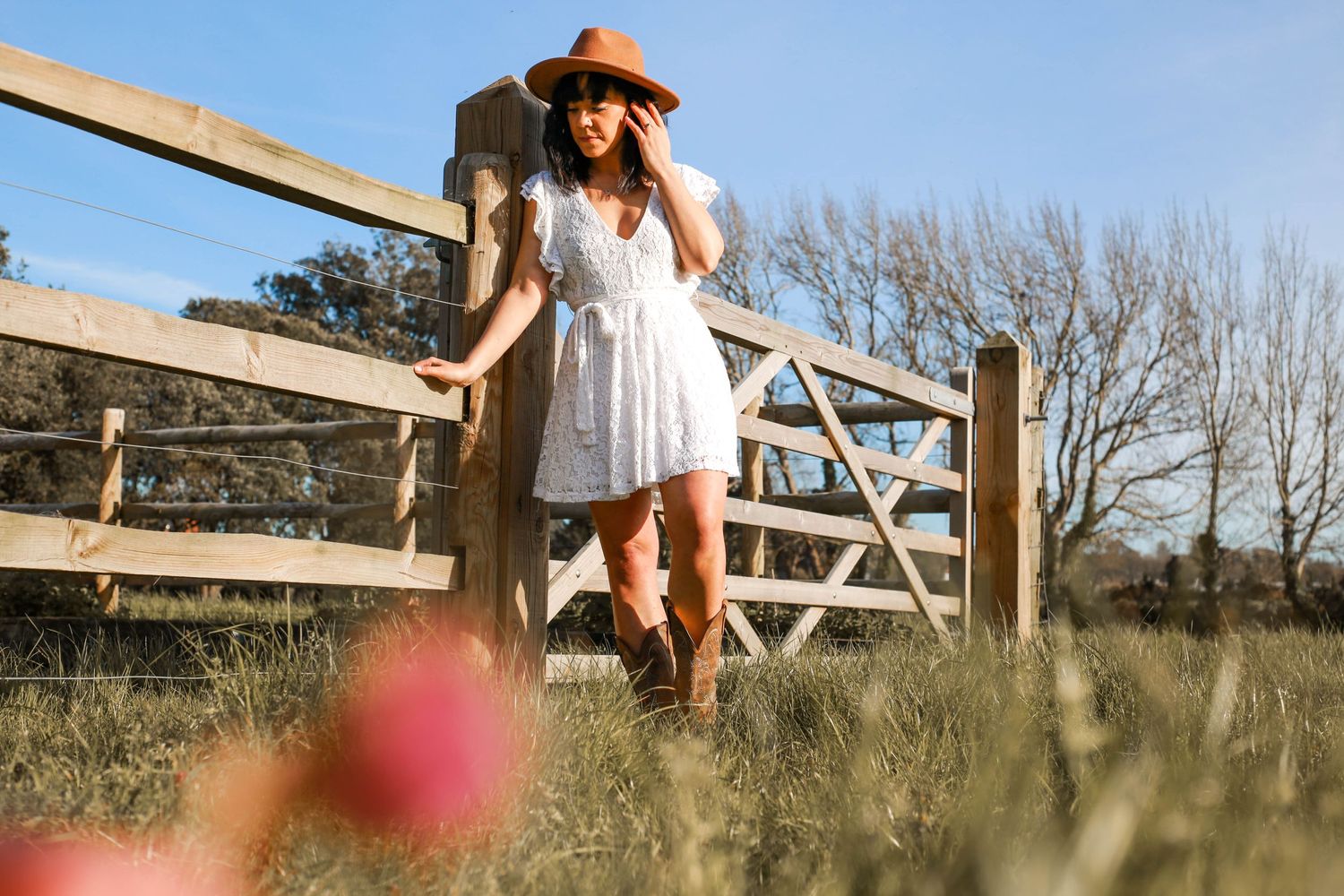 Person in white dress and hat leaning on wooden gate in rustic countryside setting.