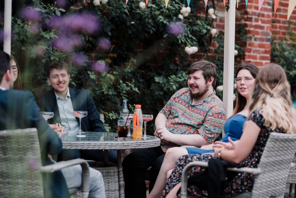 Garden party guests enjoying drinks and conversation at outdoor table.